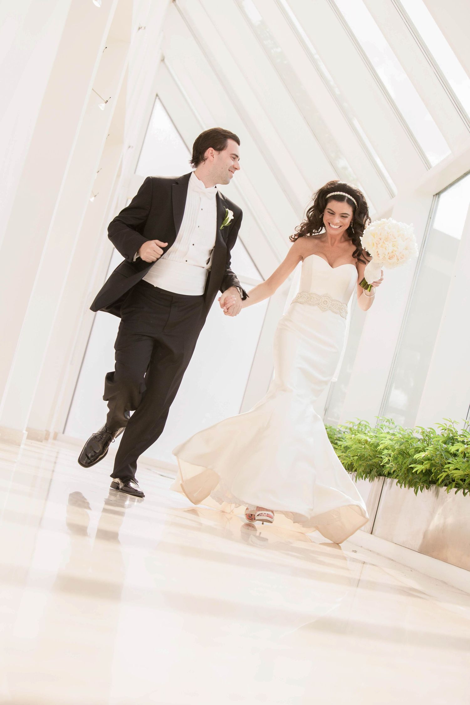 A bride and groom pose playfully in a bright modern white event space with minimalist decor and green foliage accents.