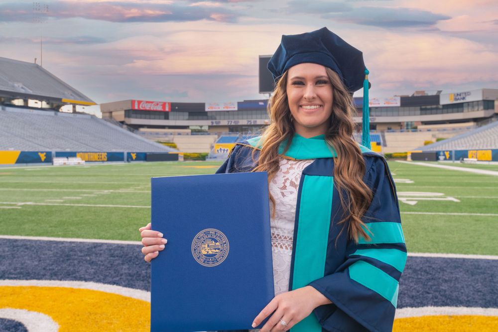 Graduate displaying diploma on football field at sunset with stadium in background.