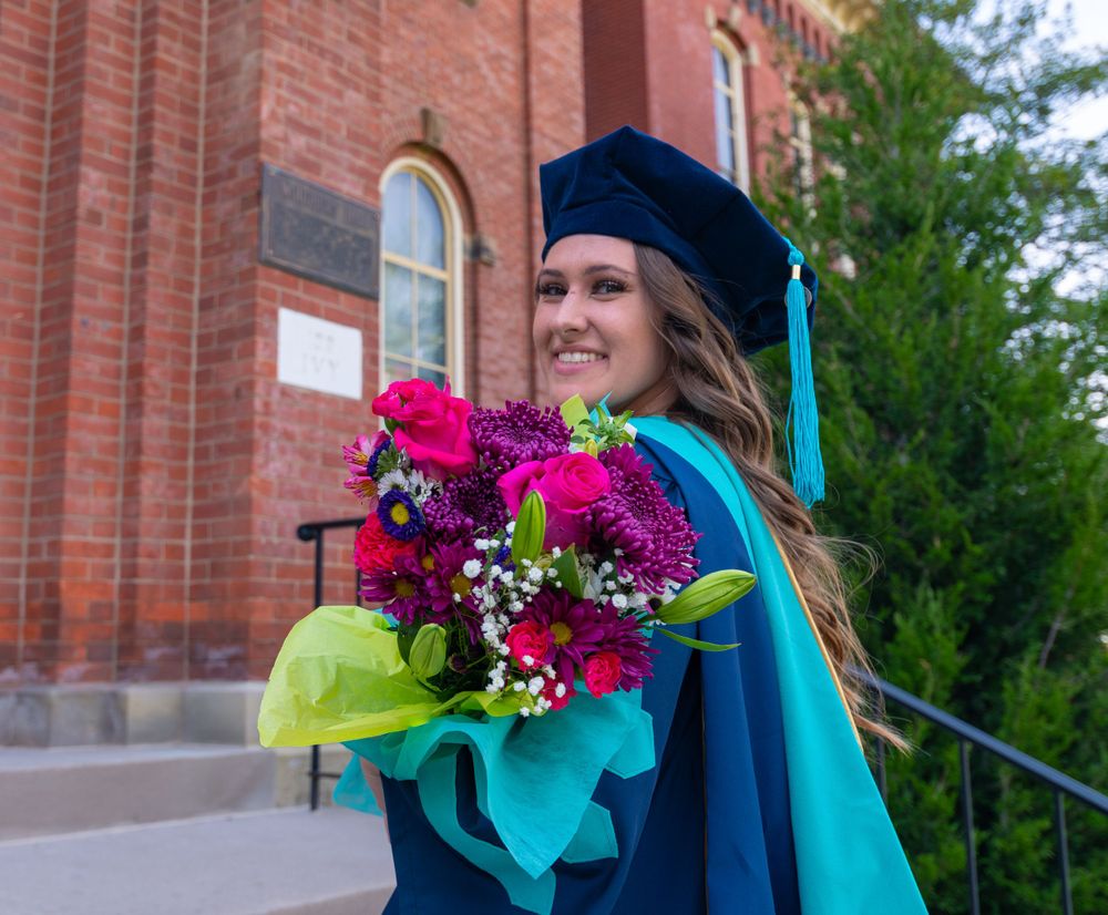 Graduate holds colorful bouquet while standing on steps of historic brick building.