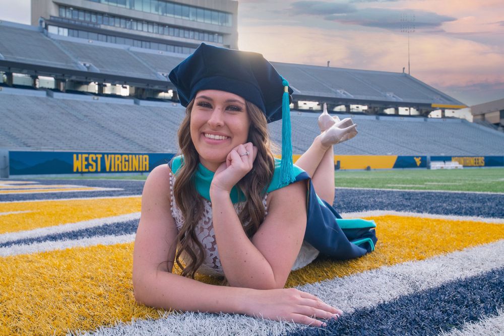 Graduate in academic regalia poses playfully on football field with stadium seating visible.