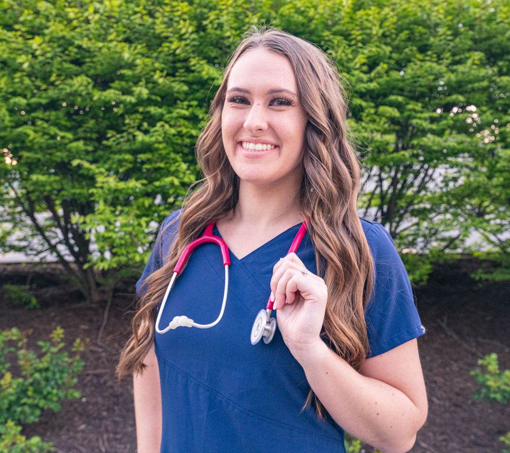 Healthcare worker in navy blue scrubs holding a stethoscope while standing in front of green foliage.