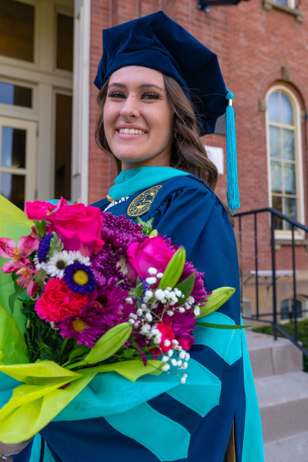 Graduate in blue doctoral regalia holds vibrant floral bouquet in front of brick building.