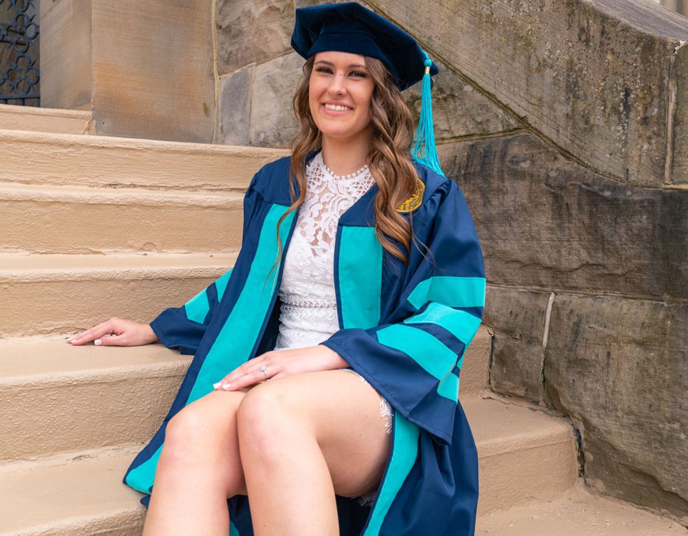 Graduate in doctoral regalia sits on stone steps wearing blue and teal graduation gown.