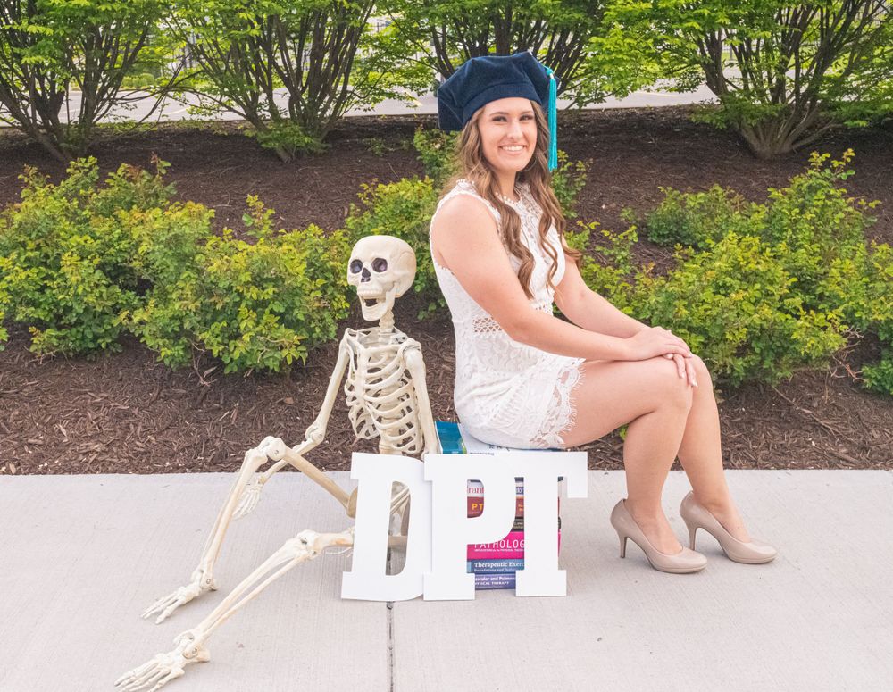 Graduate in cap poses with skeleton decoration on outdoor bench.