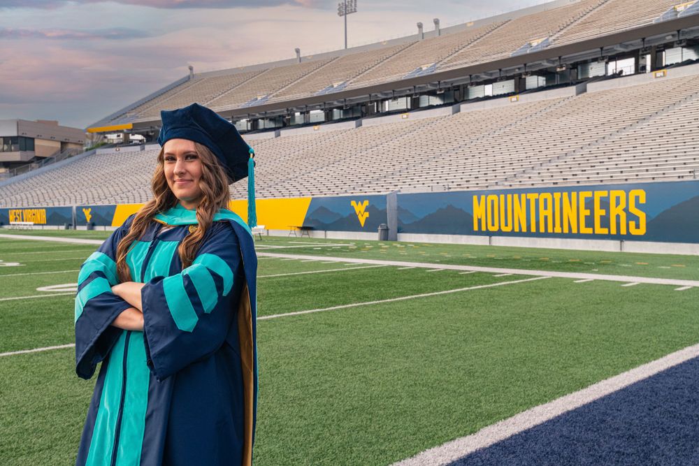Graduate in blue and teal academic regalia stands on football field with Mountaineers sign visible.