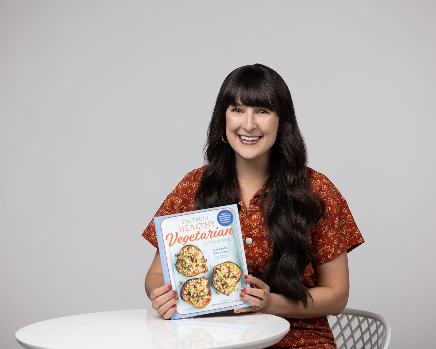 Woman in orange floral dress showcasing cookies and baked goods in a bright studio setting.