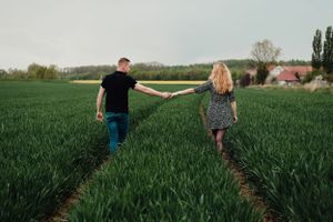 Two people walk hand in hand through a lush green agricultural field on a sunny day.