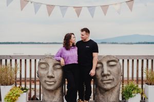 A couple poses between two large stone face sculptures near a waterfront with bunting overhead.