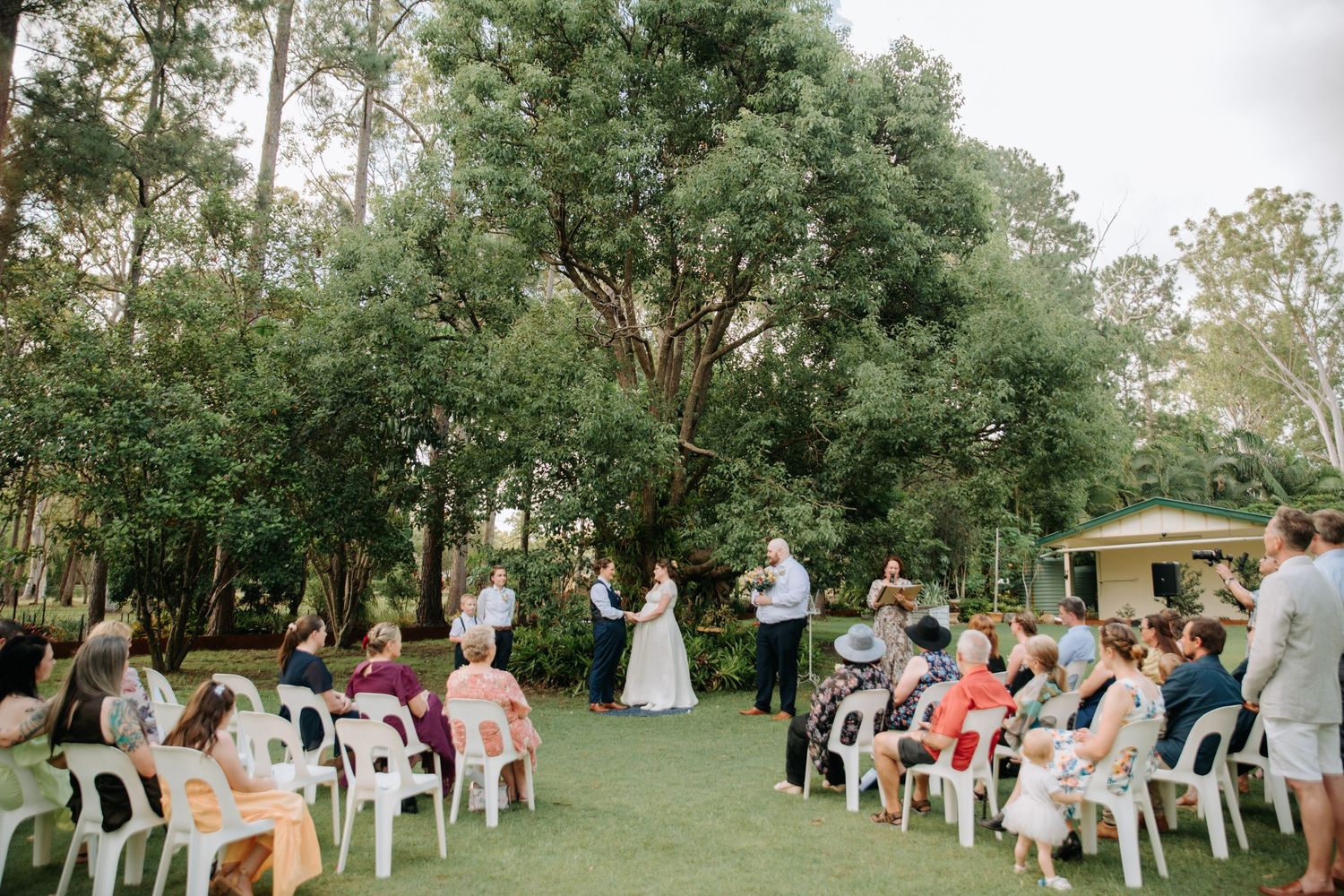 Outdoor wedding ceremony with guests seated in white chairs surrounded by tall trees.