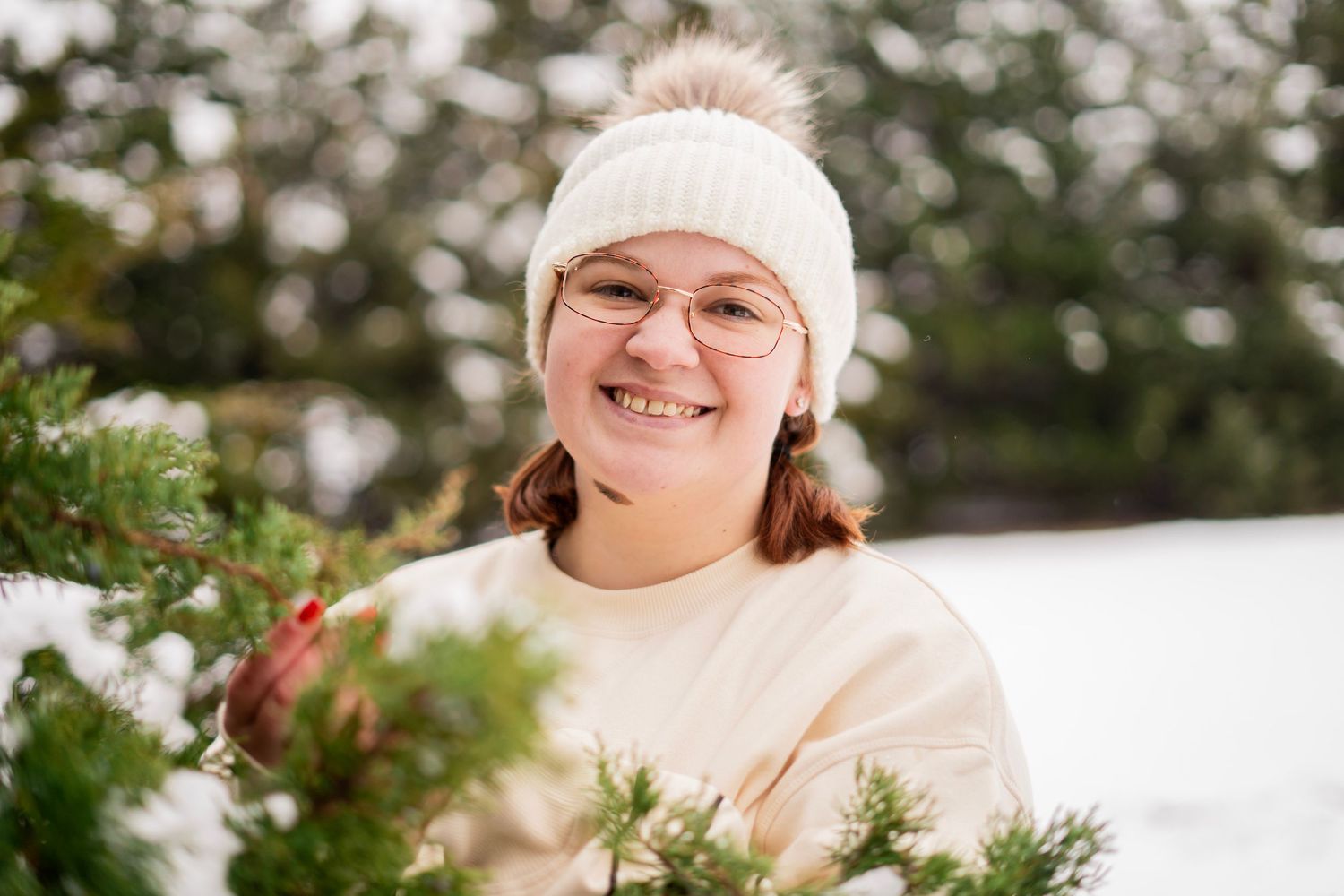 Happy young woman wearing glasses and white knit hat smiling beside snow-dusted pine branches in winter.