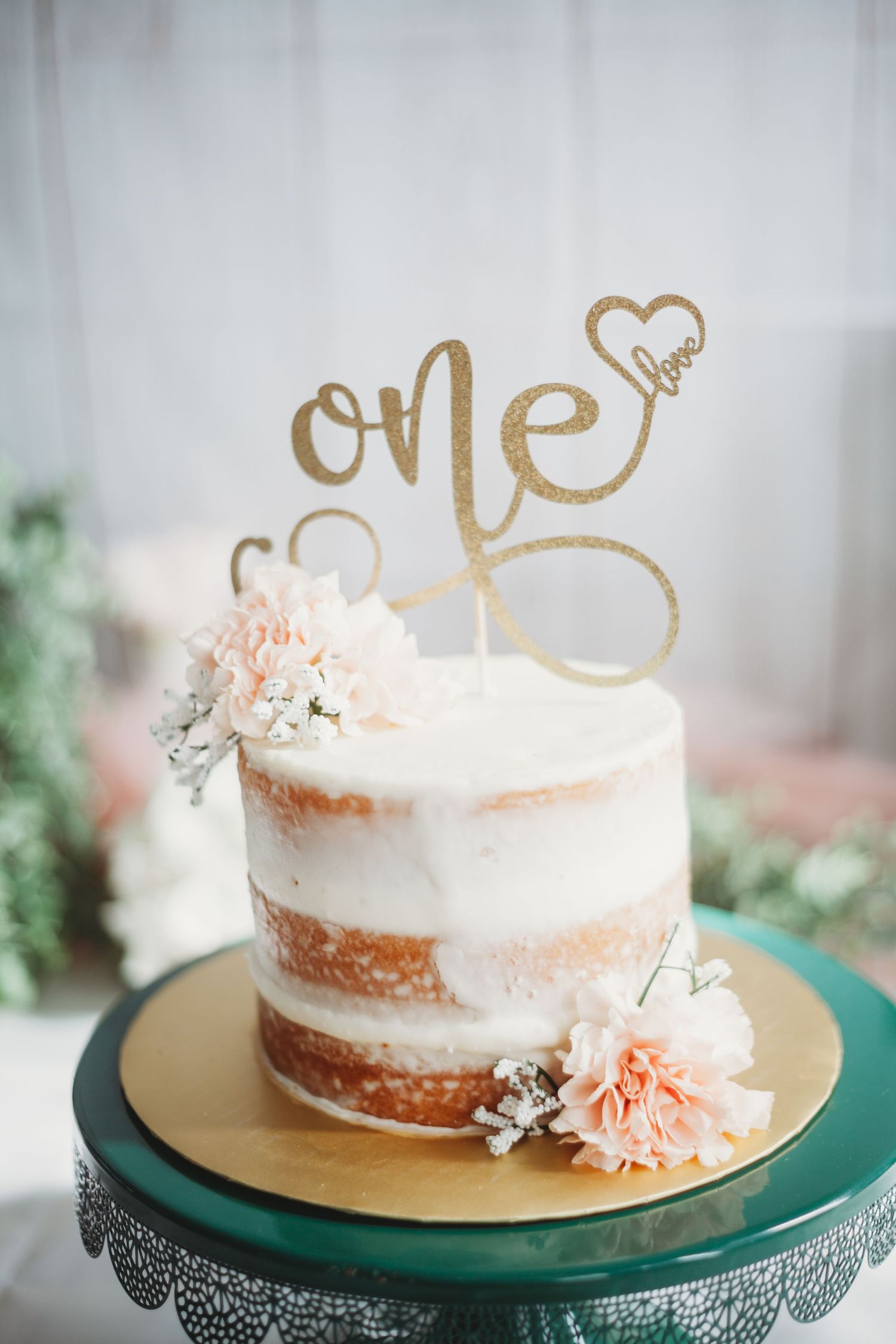 Elegant semi-naked wedding cake with gold topper and peach floral accents on green stand.