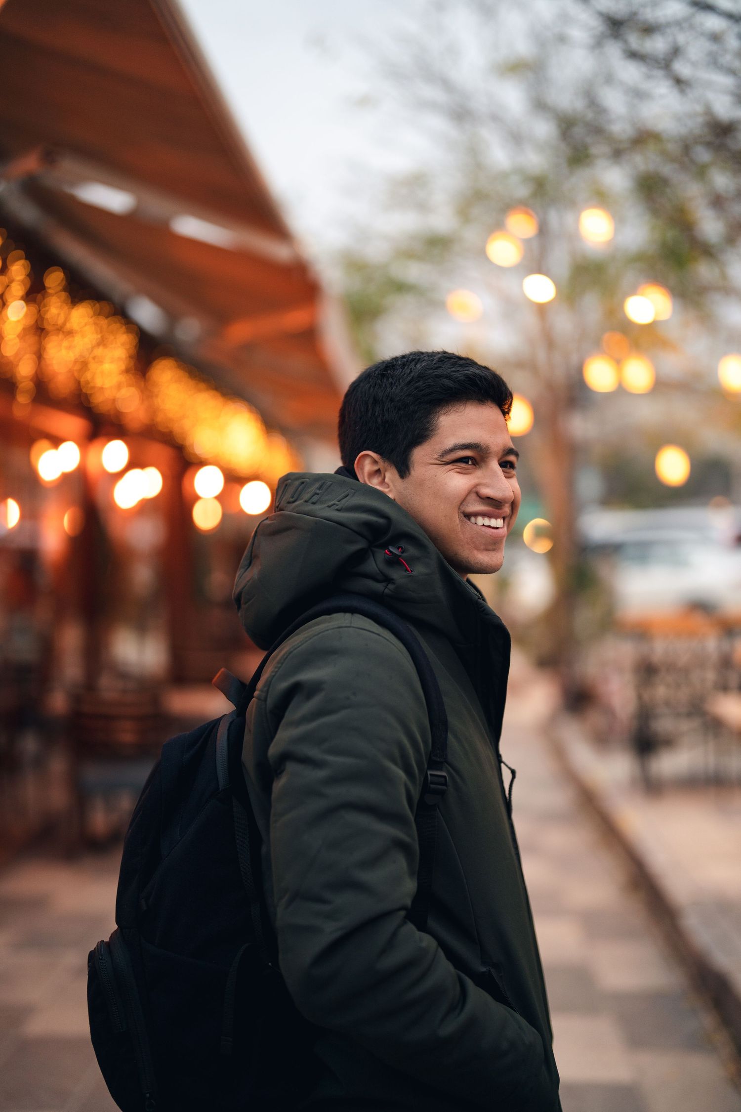 Person in dark coat smiling outdoors with warm evening lights in background.