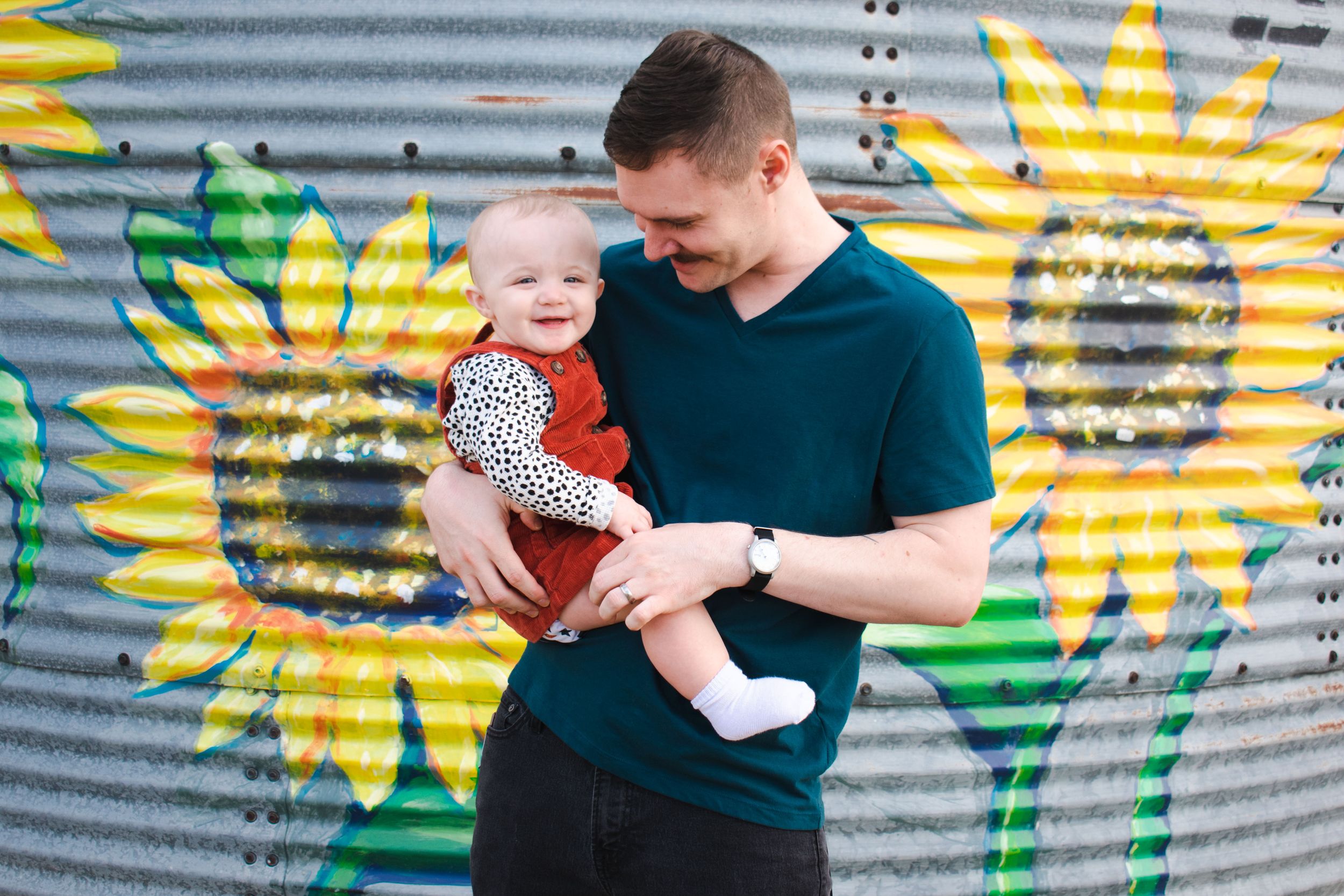 A young father smiles down at his baby girl as he poses in front of a floral  mural