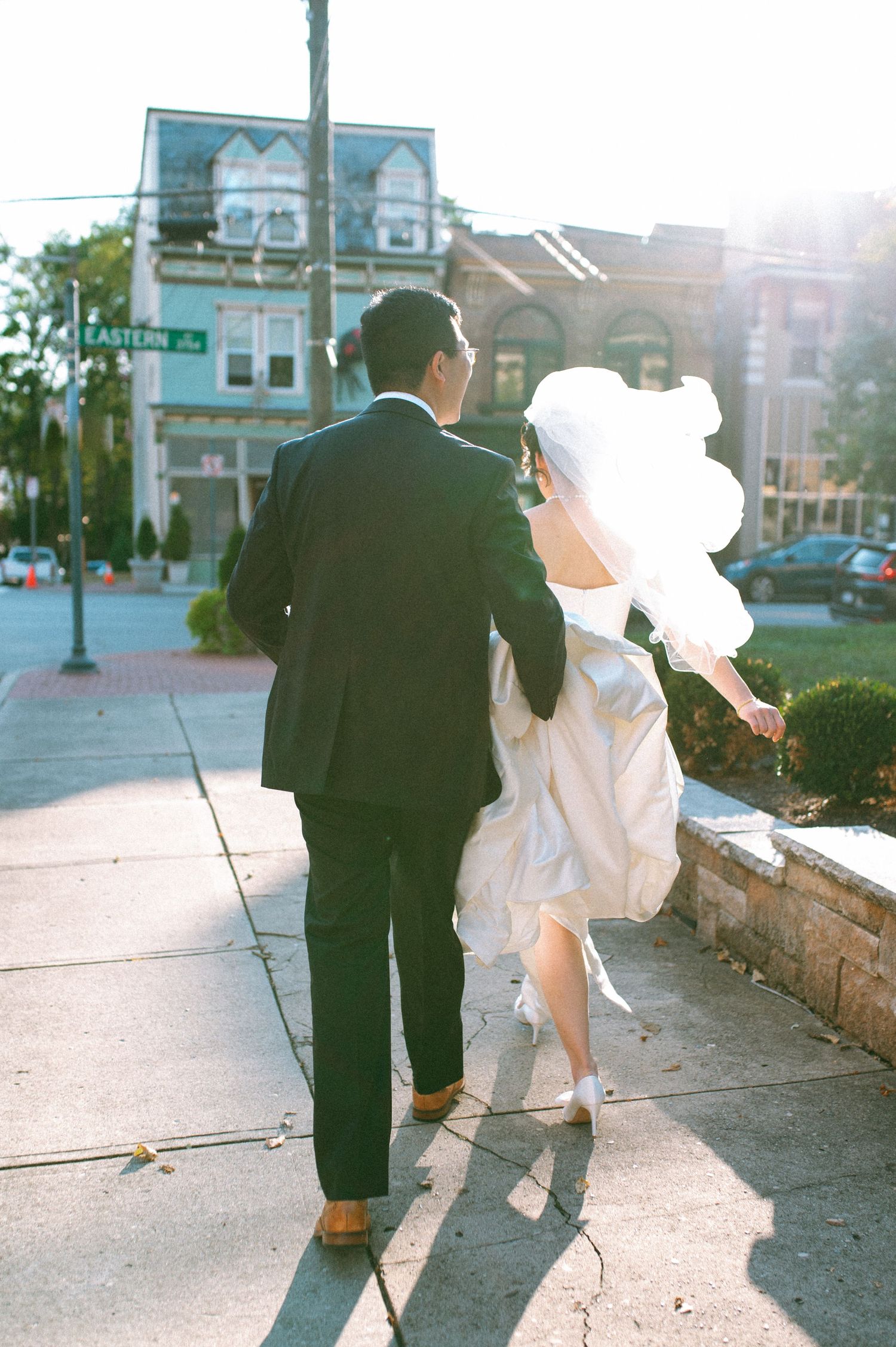 Newlywed couple walking down sunny sidewalk in formal wedding attire.