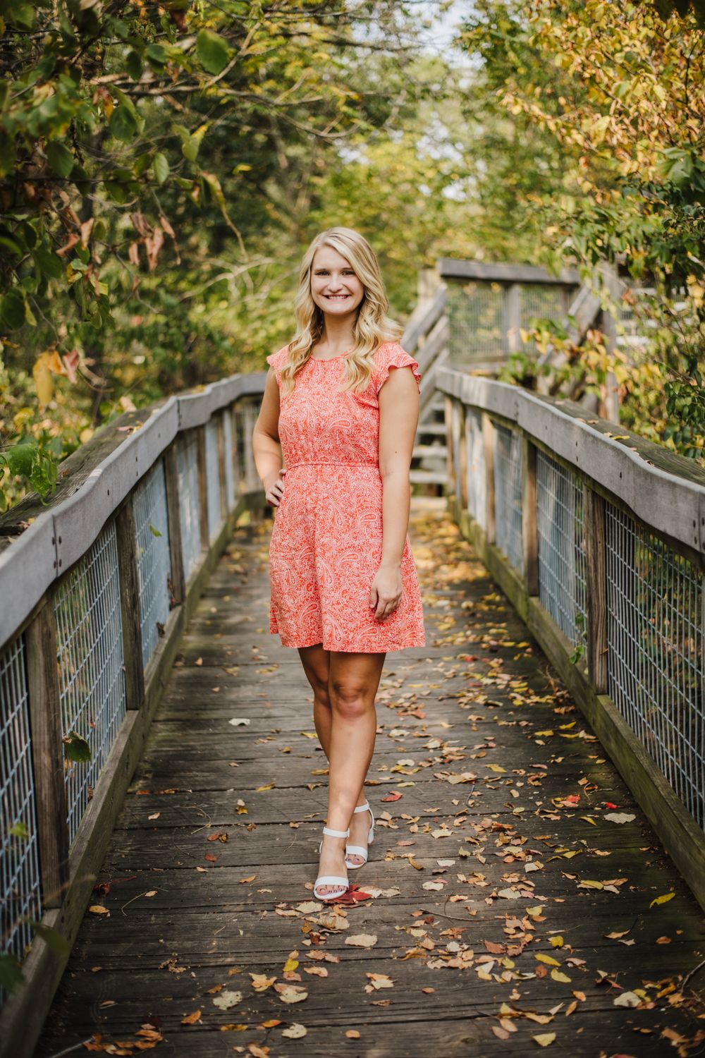Walking along wooden boardwalk surrounded by autumn foliage wearing coral dress.