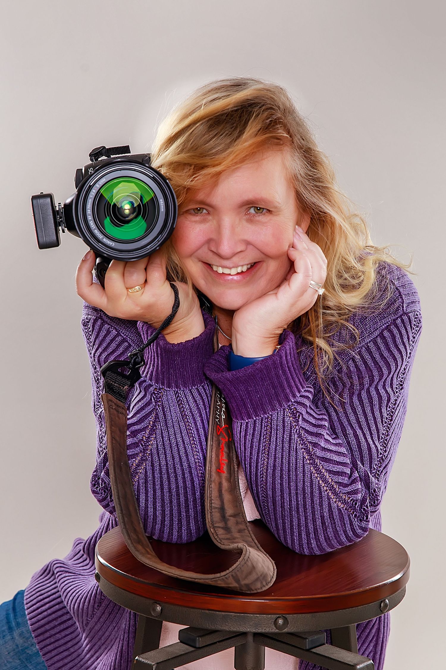 Photographer holding DSLR camera while wearing purple sweater in studio portrait.