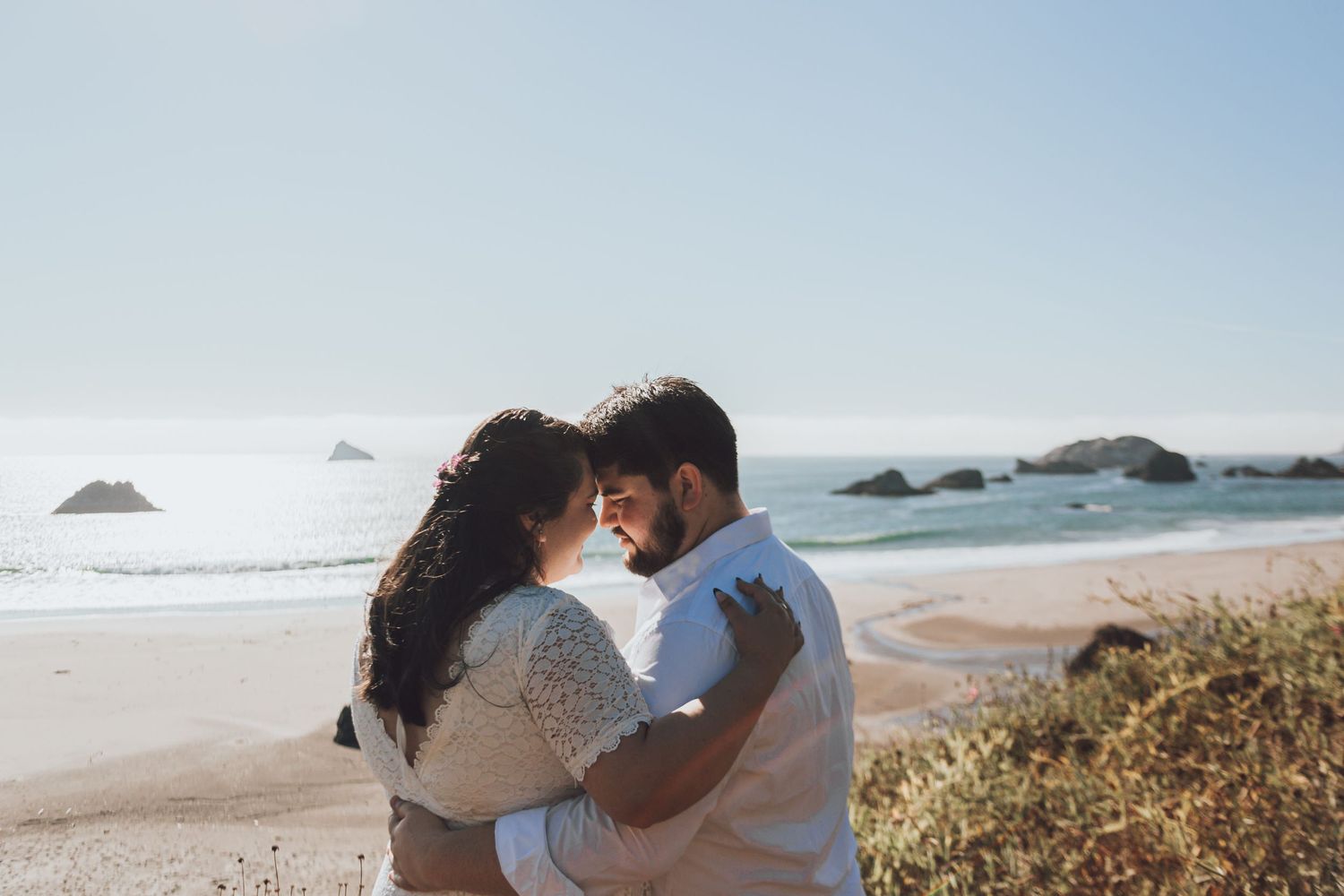 A romantic moment between a couple embracing on a sunny beach with coastal rocks in the background.