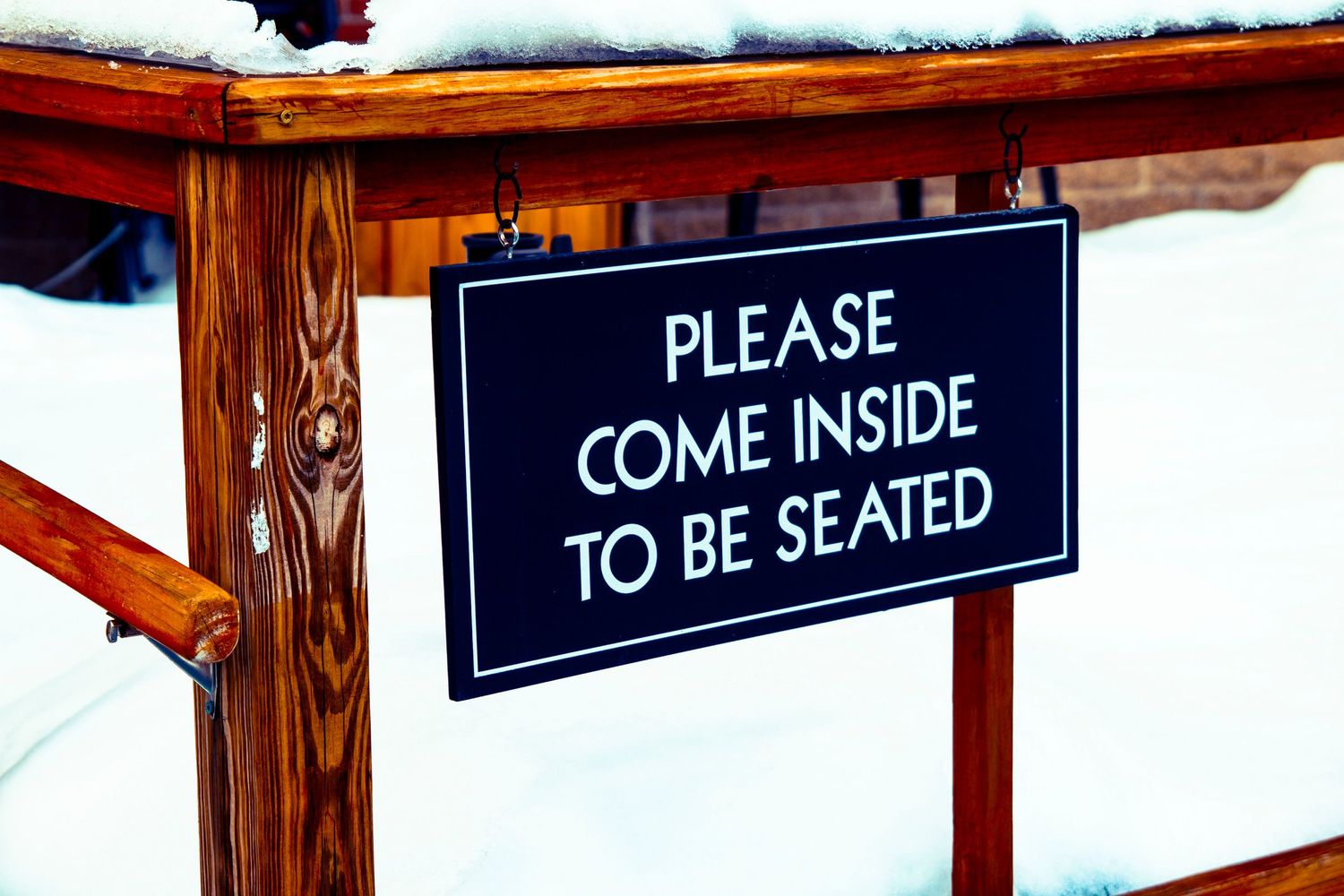 A wooden sign reads 'Please Come Inside To Be Seated' mounted on a snow-covered surface.