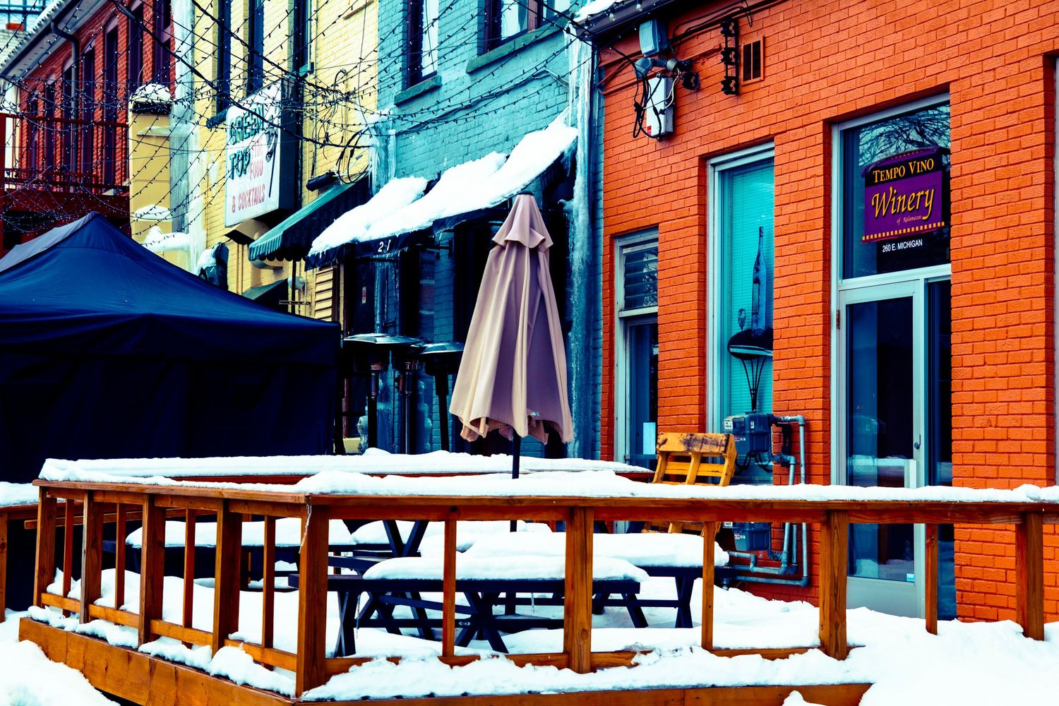 Colorful urban street scene with snow-covered outdoor seating and bright building facades.