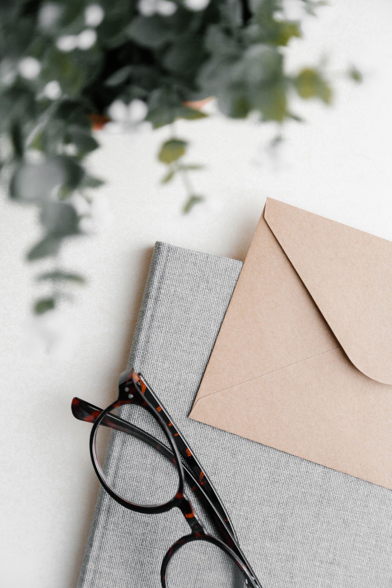 Minimalist flatlay with reading glasses, grey book and beige envelope against white background.