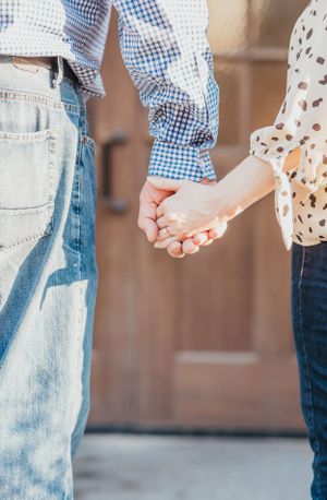 People holding hands wearing blue gingham shirt and floral print top against brick background.