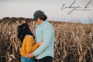 A couple embraces in front of dried corn stalks during a sunset photoshoot wearing yellow and light blue.