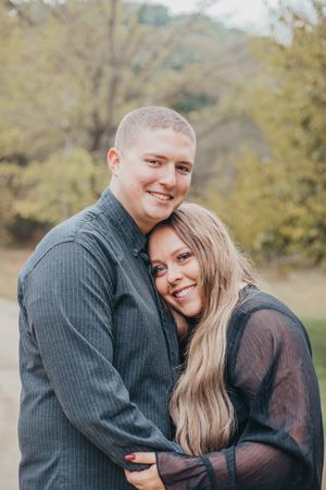 A couple embraces outdoors in autumn, wearing dark sweaters against a blurred natural background.