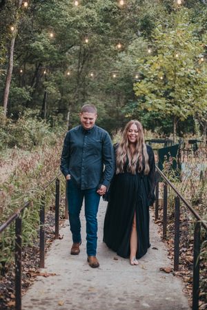 A couple walks hand in hand on a garden path with string lights overhead and autumn foliage in the background.
