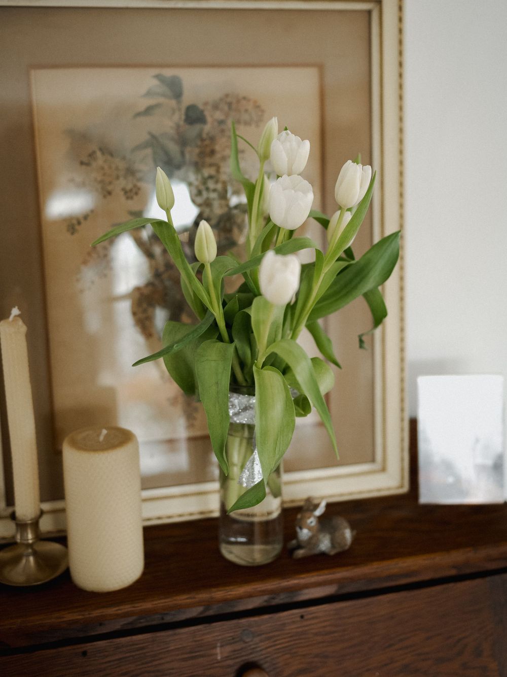 White tulips in a glass vase displayed on a wooden shelf with framed artwork.