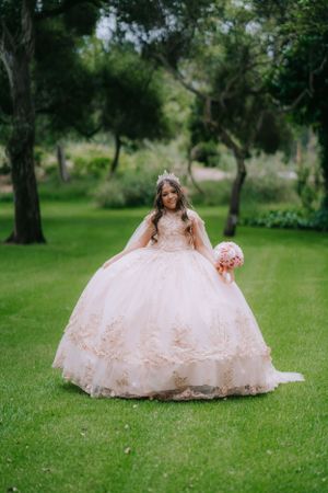 A young woman in a voluminous blush quinceanera gown poses in a lush garden setting.
