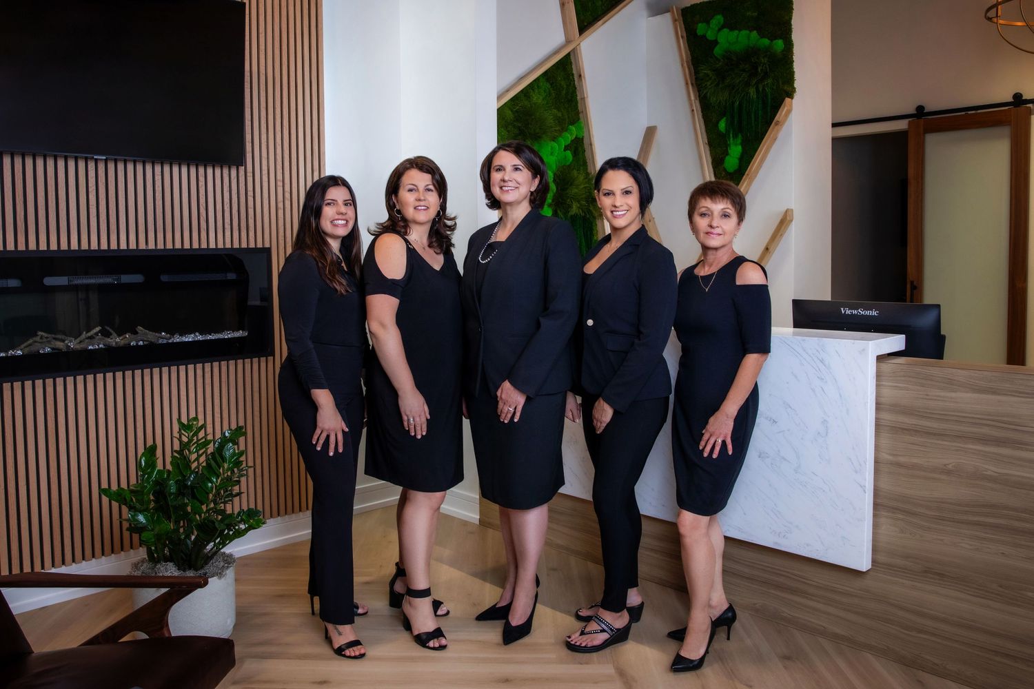 Group of five women in black dresses posing in modern office setting.