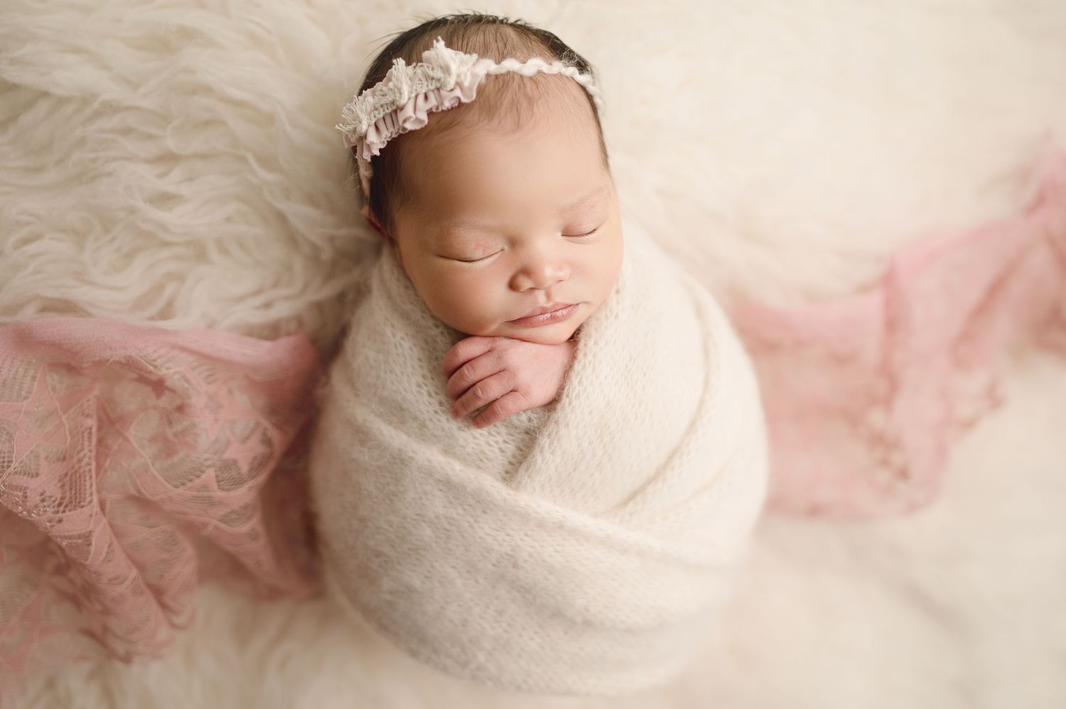 Sleeping newborn wrapped in white swaddle wearing delicate lace headband on cream backdrop.