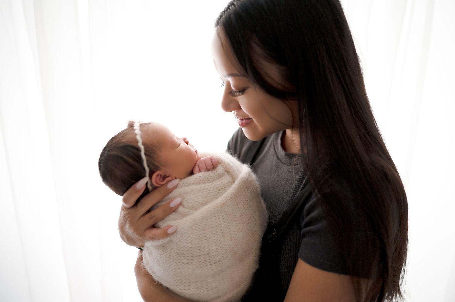 Mother in dark shirt tenderly holds swaddled baby in bright window light.