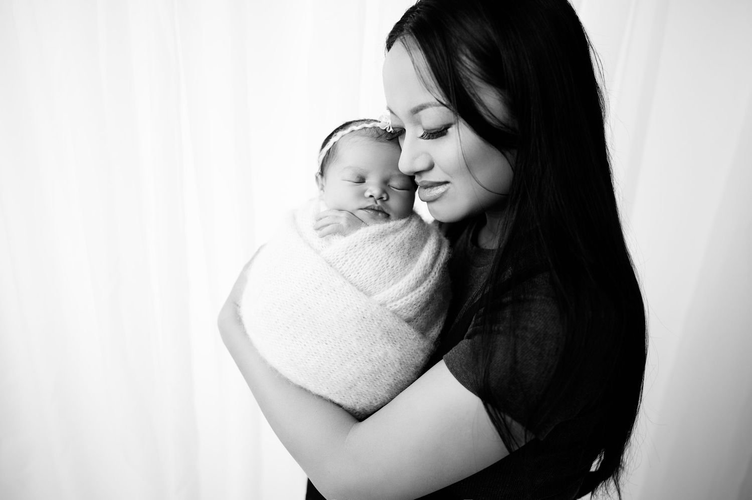 Black and white portrait of mother holding wrapped newborn against bright backdrop.