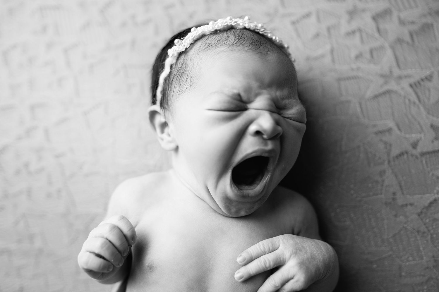 A black and white close-up photo of a yawning baby wearing a headband.