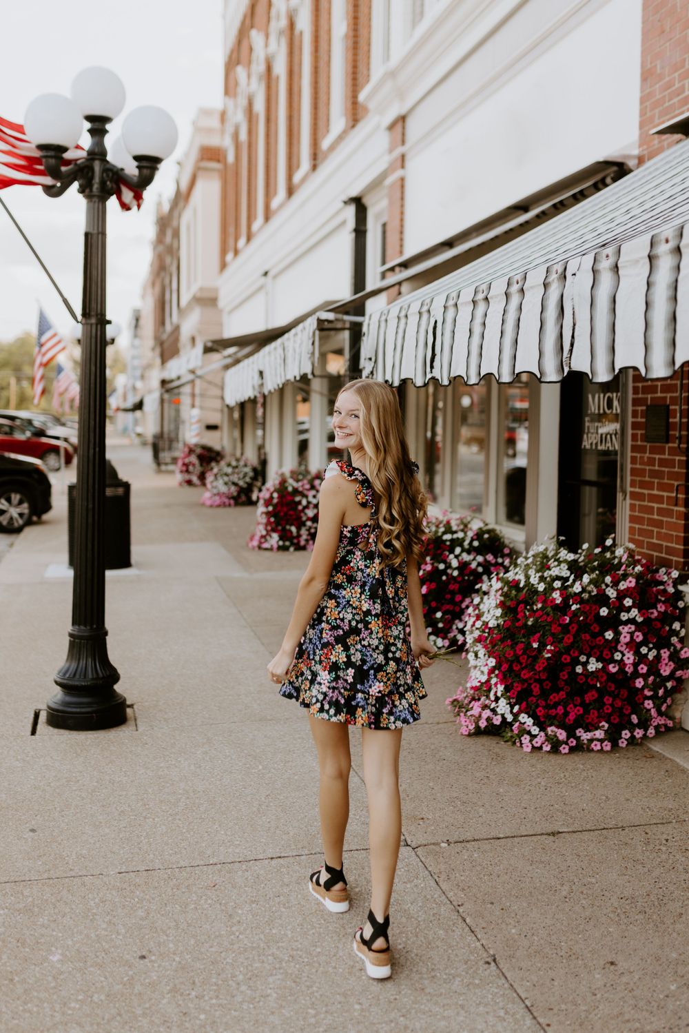 Young woman in floral dress walking down a charming small-town street with flowers and lampposts.
