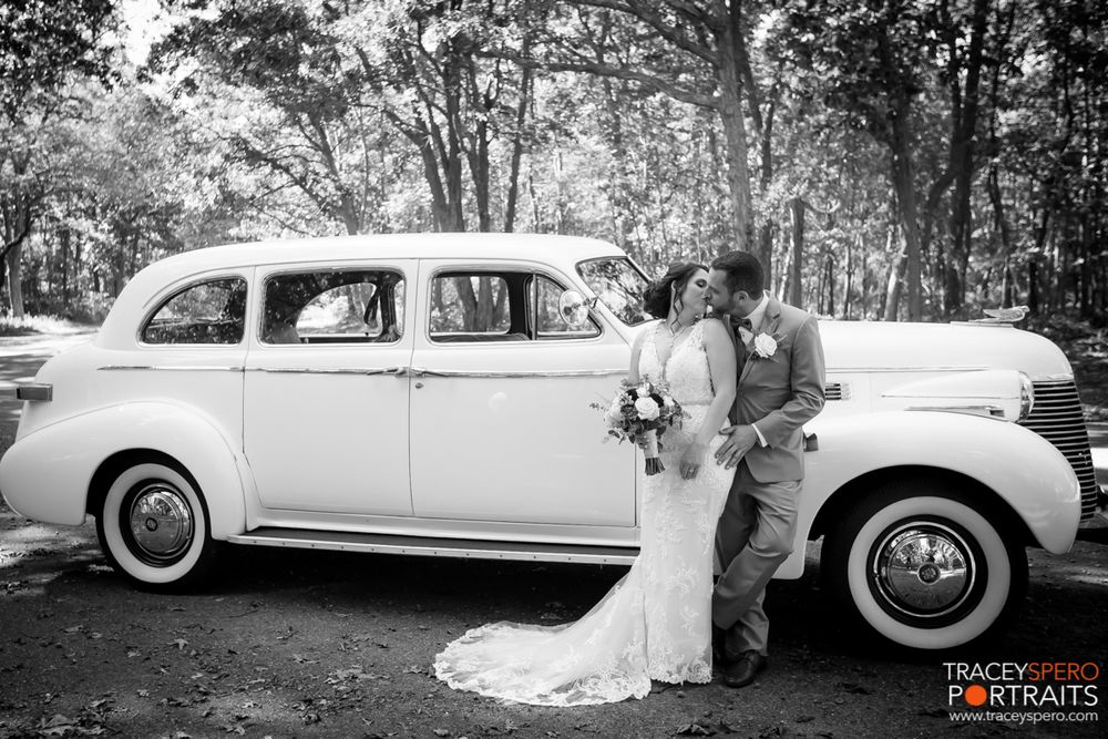 Classic black and white photo of newlyweds posing beside vintage car.