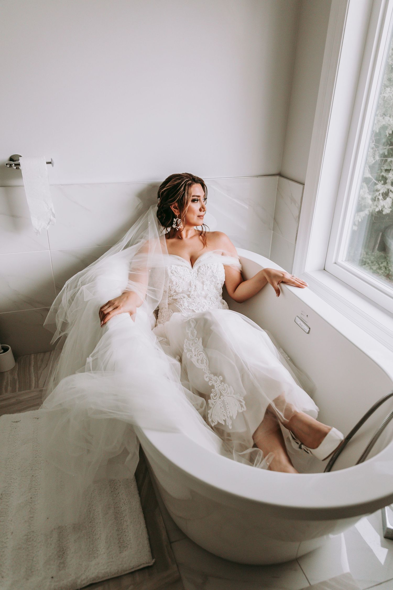 A bride in a white lace gown poses elegantly in a vintage bathtub near a window.