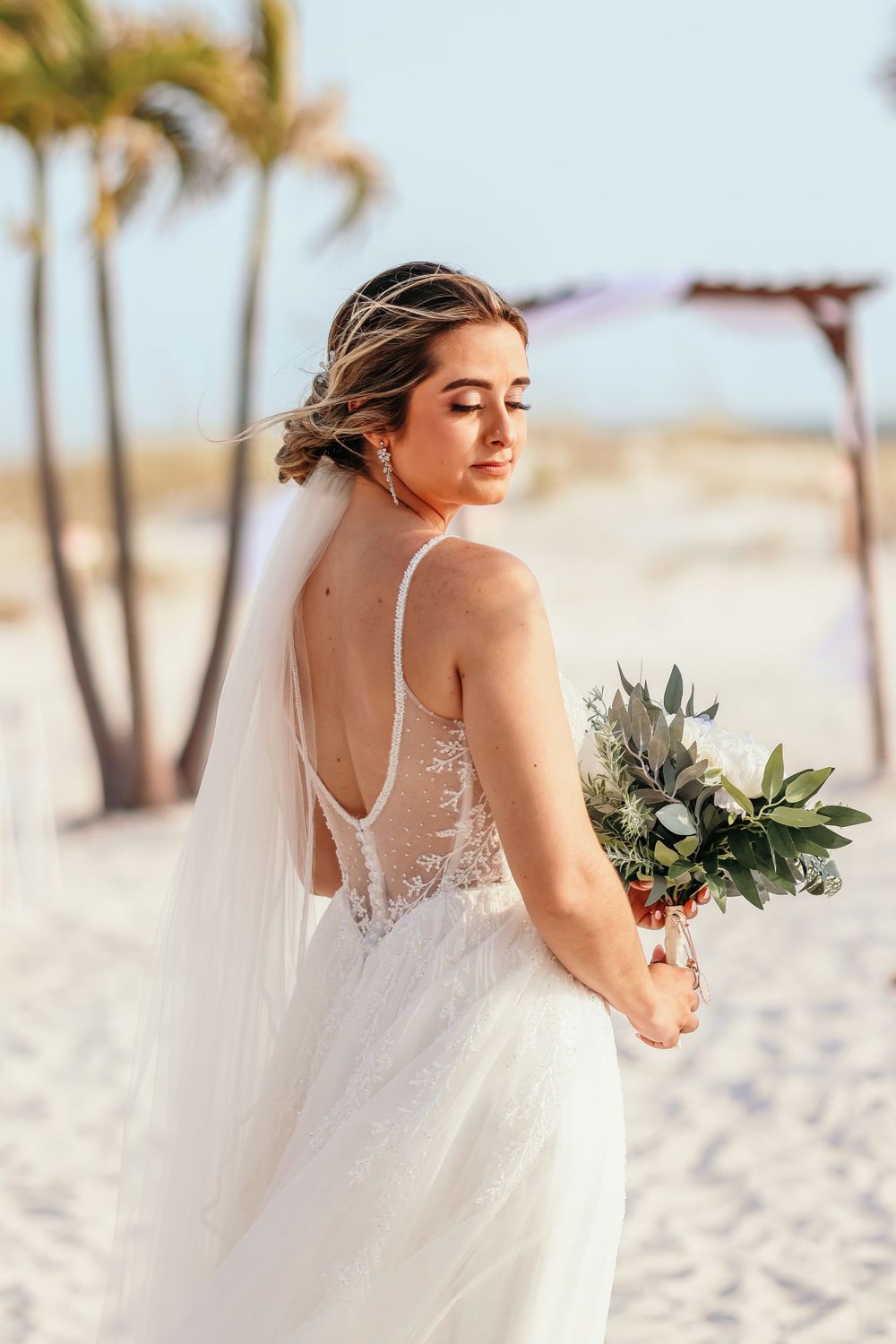 Bride in a flowing white dress poses gracefully on a sunny beach with palm trees.