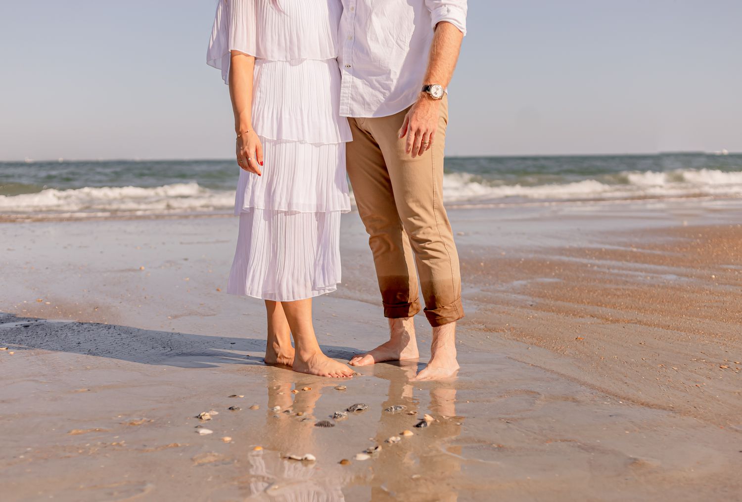 Barefoot couple standing together on sandy beach with ocean waves in background.