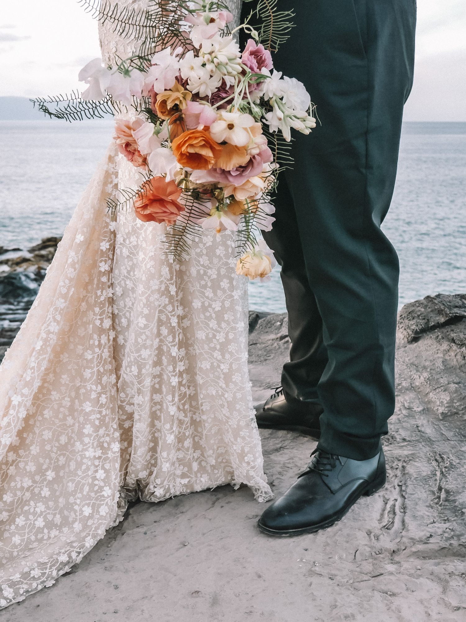 A white lace wedding dress with peach and pink floral bouquet details photographed against an oceanside backdrop.