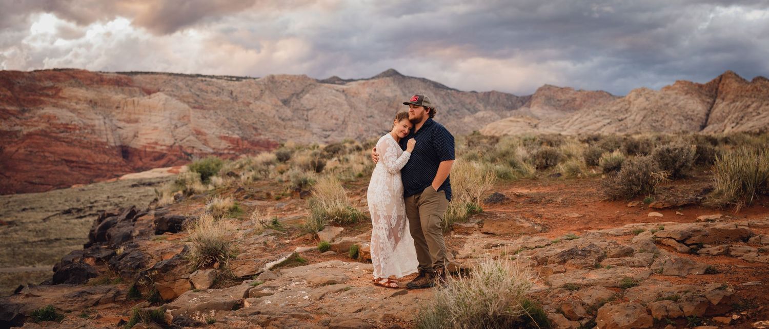 Couple sharing a romantic kiss on red desert terrain with stormy purple sky and red rock mountains behind them.