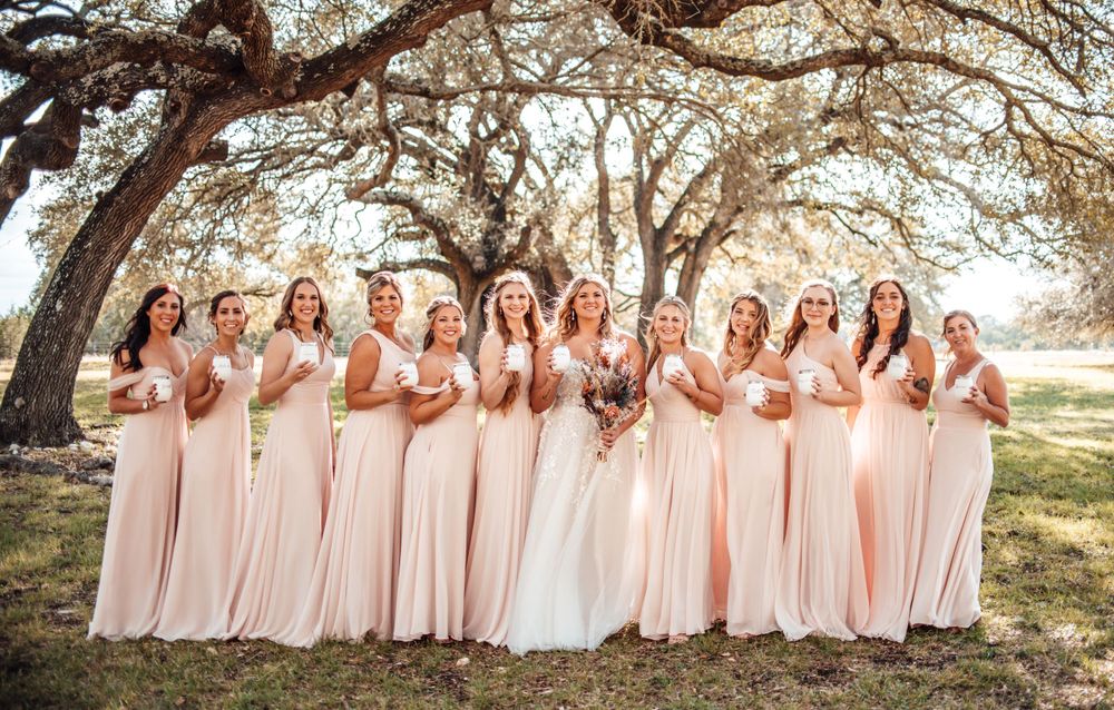 Bridesmaids in matching blush gowns pose together under a sprawling oak tree.
