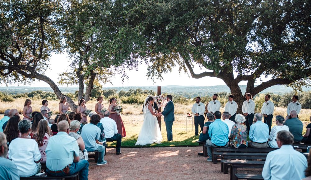 Outdoor wedding ceremony under sprawling oak trees with guests seated on wooden benches.