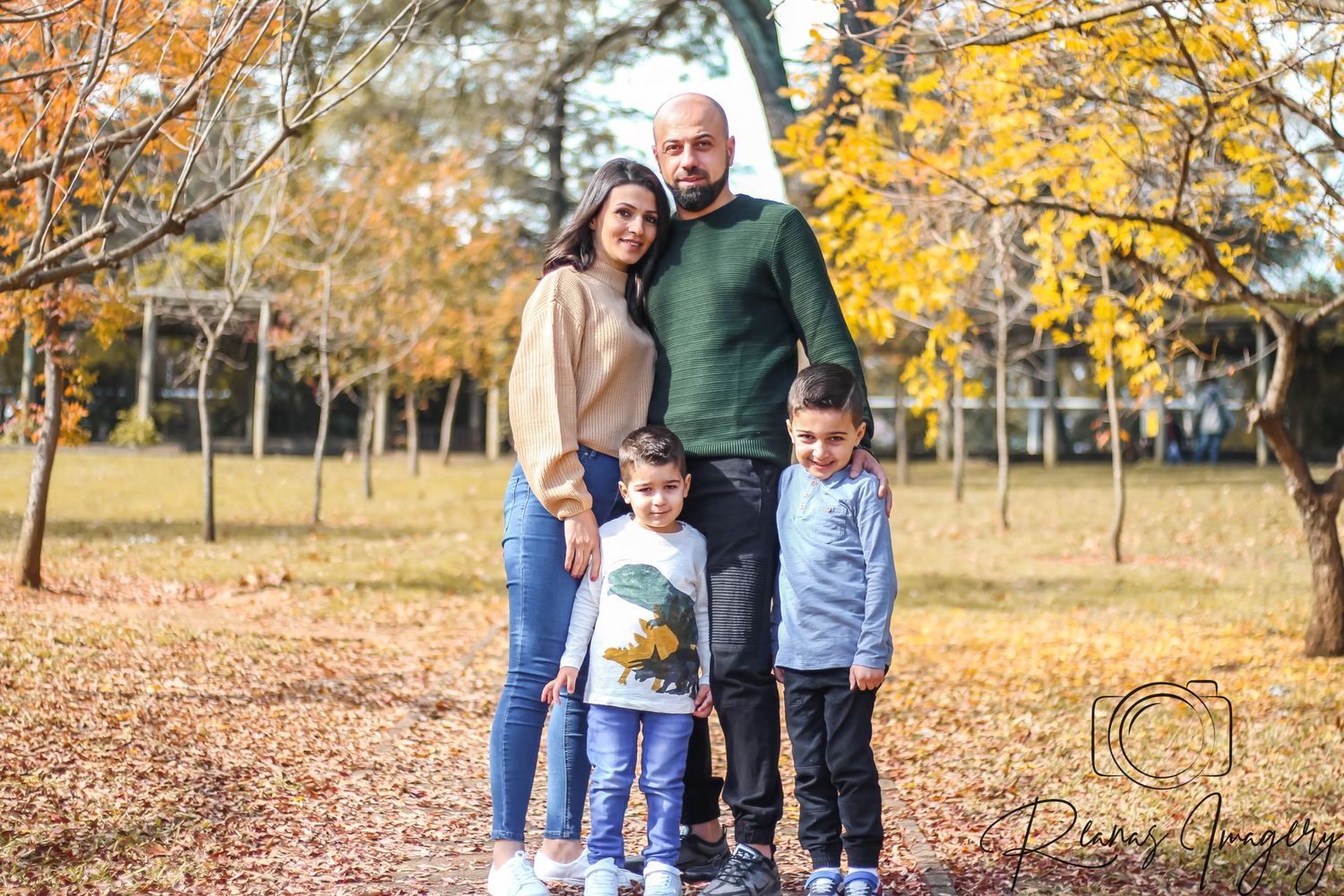 Family of four with two young boys posing together outdoors among colorful autumn foliage.