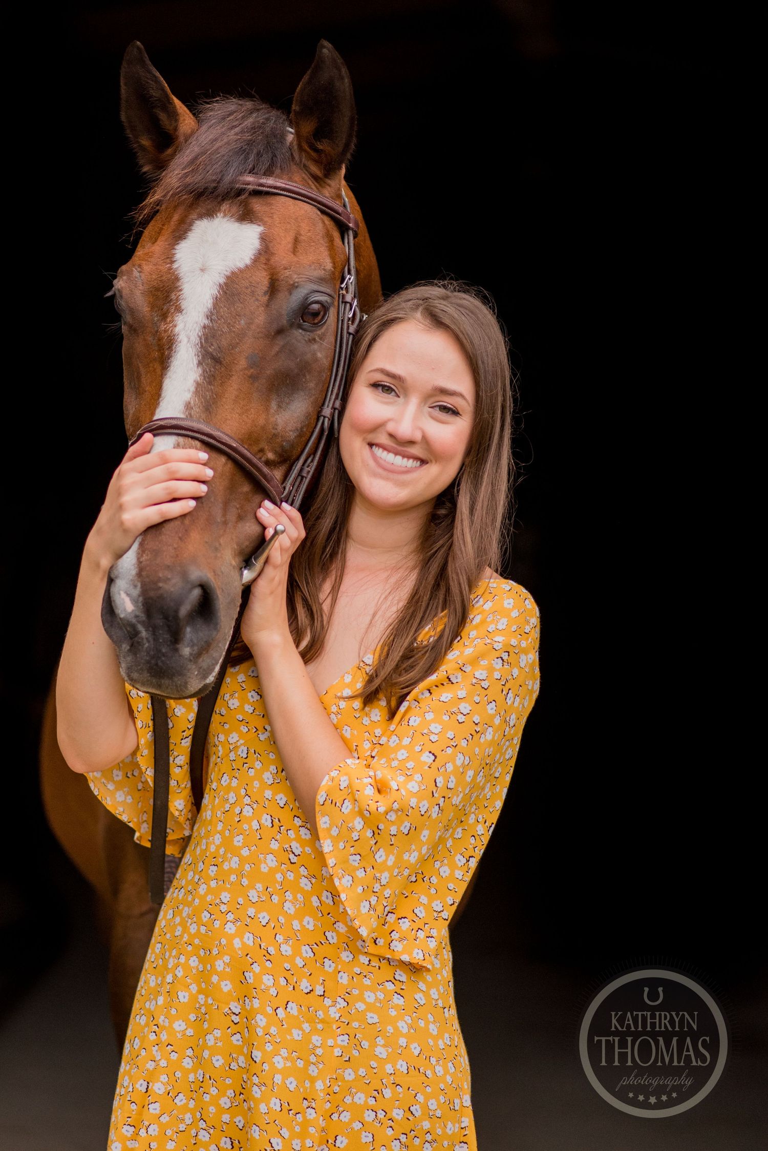 A beautiful bay horse with white blaze poses with someone in a yellow floral dress against dark background.
