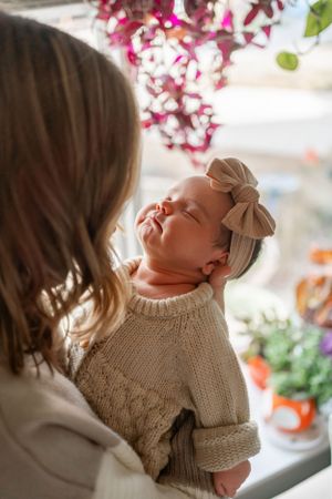 A baby wearing a knit sweater and bow stretches near blooming pink flowers.