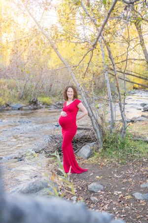Expecting mother in flowing red dress poses by stream at sunset.