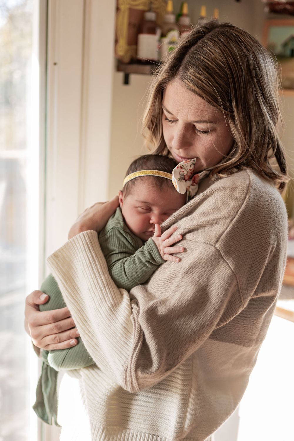 Tender moment between parent and sleeping infant by sunlit window.
