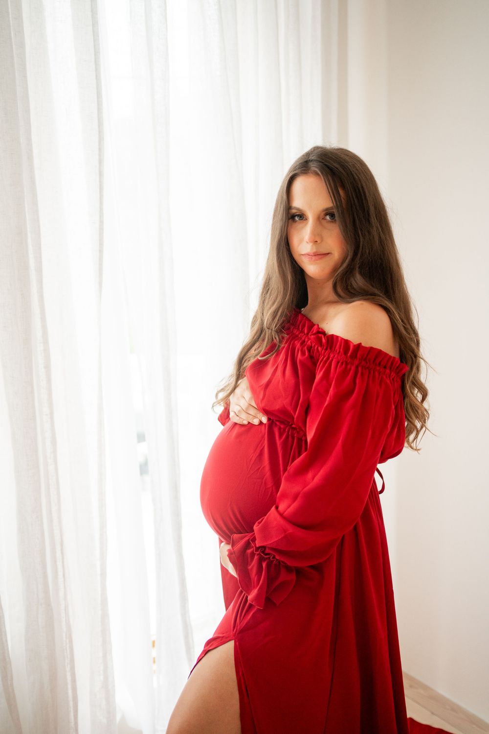 An off-shoulder red maternity dress with long sleeves photographed against white curtains.